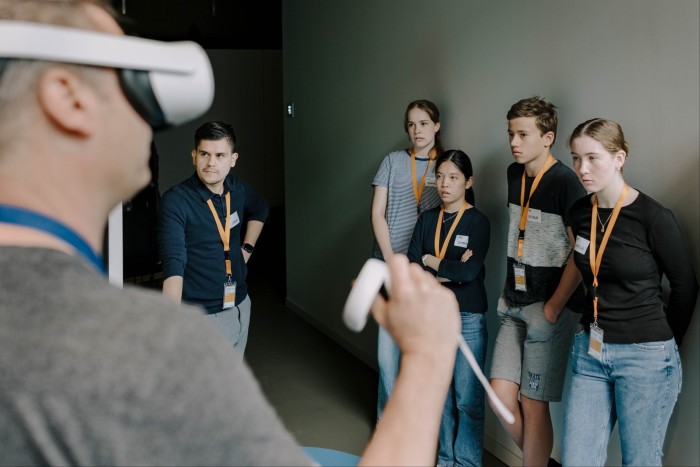 A group of young adults stand attentively watching a man wearing a virtual reality headset