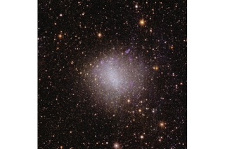 A glittering pink-white blob of light in the center of the image, surrounded by millions of spots of light representing distant cosmic objects.