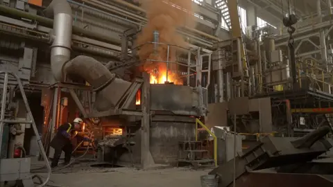 BBC News / Kevin Church Flames rising from the top of the arc furnace as material is added to the molten steel