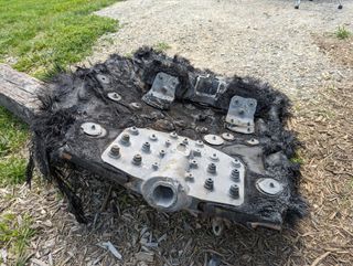 A large black piece of fiberglass covered with metal bolts and plates lies on the ground next to a path leading into a forest. Mountains can be seen in the distance